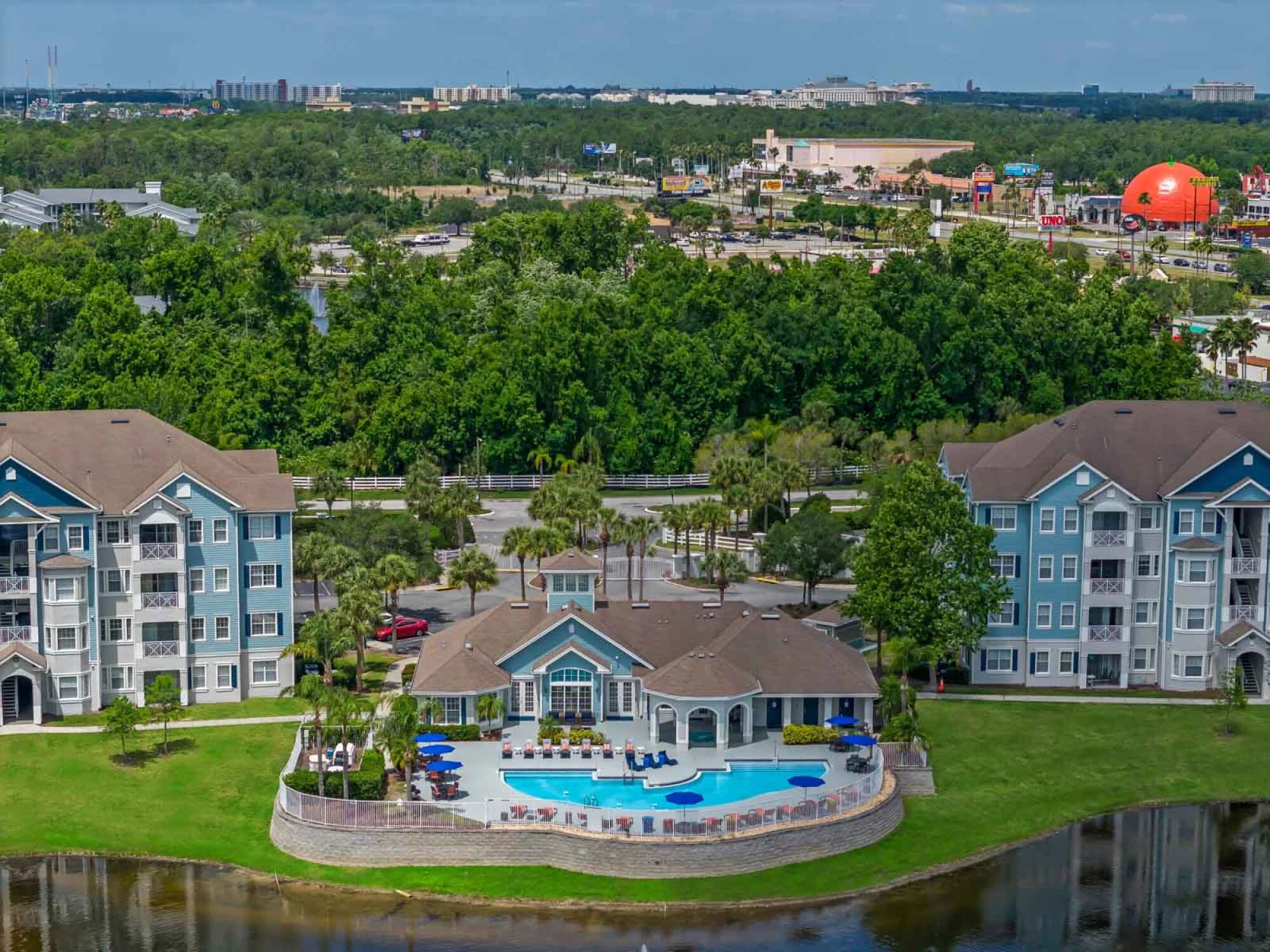 An aerial view of the property with the pool and office building in the center with four-story blue and white buildings on either side showing trees and area attractions.